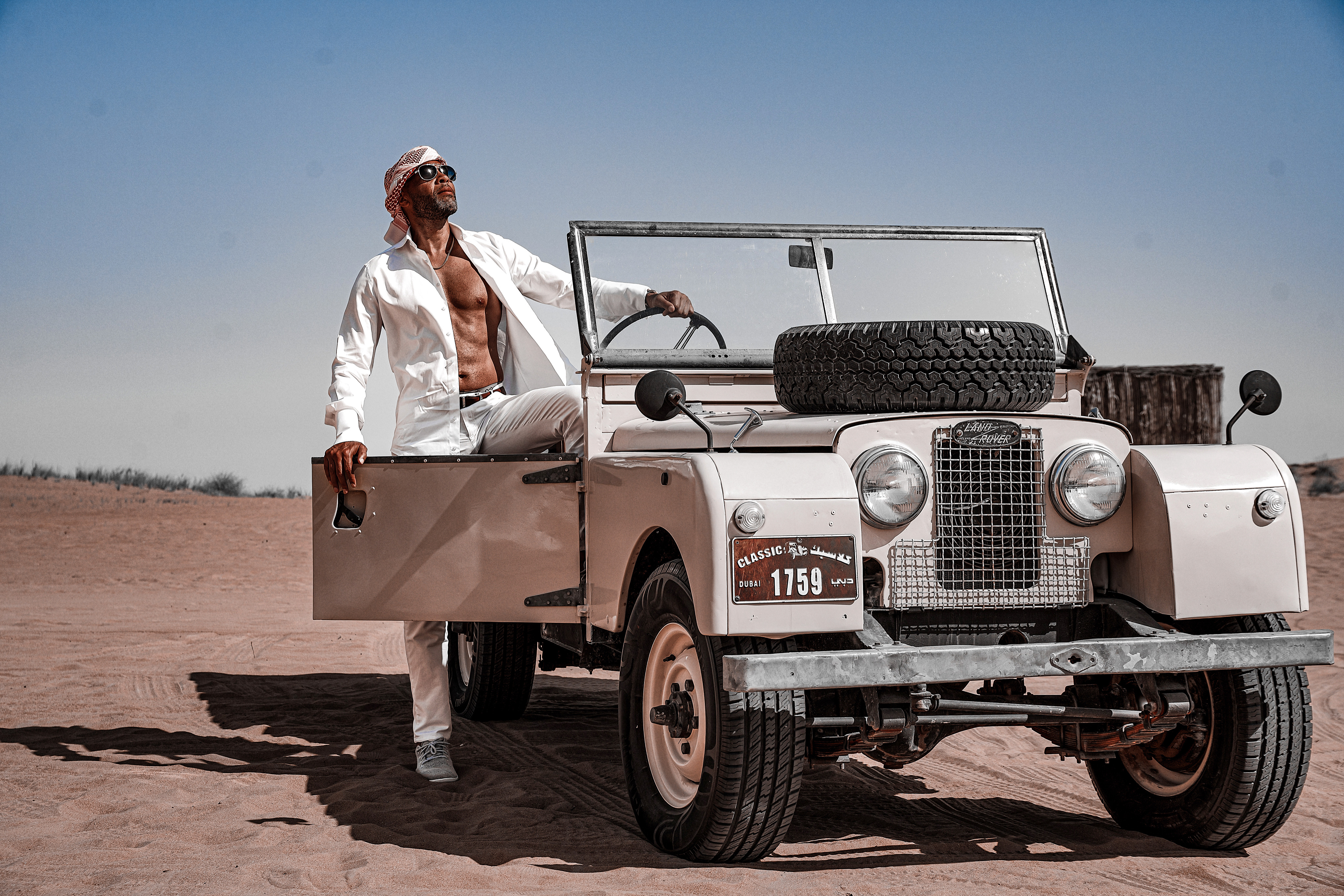In the desert, a man is striking a pose next to a vintage jeep during a luxury car photoshoot in Dubai.